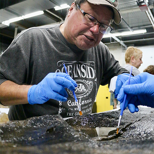 A man in grey shirt and grey ball cap works to begin the preservation process of the canoe.